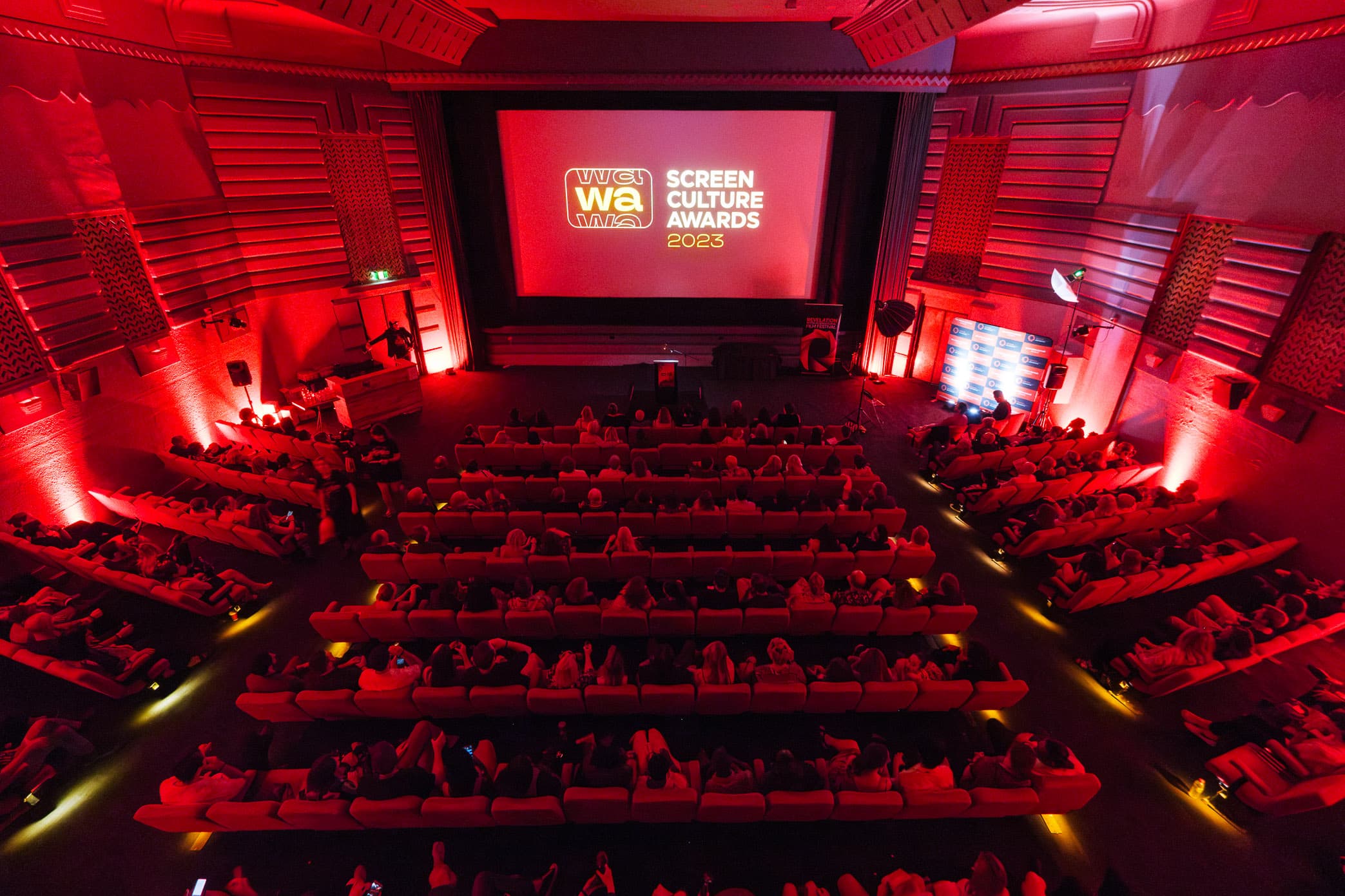A theater filled with people watching a screen displaying "Screen Culture Awards 2023" with a red-lit ambiance.
